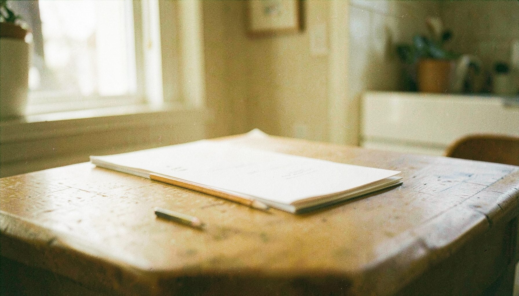 A grainy analog film photograph of a wooden kitchen table with a sharpened pencil and a neat stack of papers, soft natural light from a nearby window.